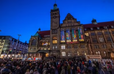Zu sehen ist das Rathaus, an dem die Friedensbanner hängen. Vor dem Rathaus steht eine Bühne auf der zwei Menschen sprechen und eiiner Bläser-Band sitzt. Vor der Bühne stehen mehrere hundert Menschen.