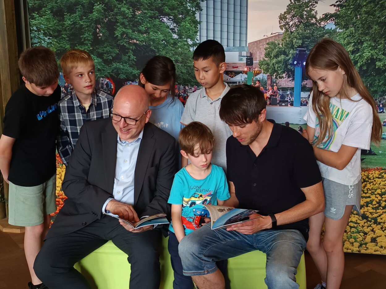 Der Oberbürgermeister hält sitzend ein Heft in der Hand. Neben ihm sitzt ein kleiner Junge und ein Mann. Hinter ihm stehen Kinder und schauen in das Heft.