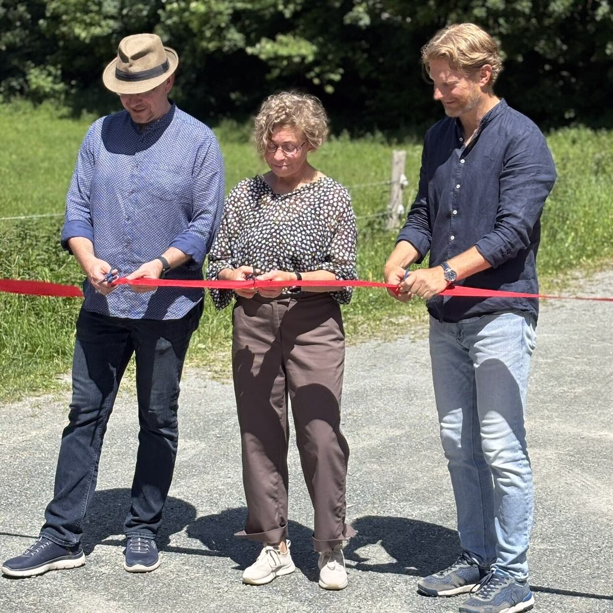 Ein Mann mit Hut, eine Frau und noch ein Mann, alle in legerer Kleidung, schneiden ein rotes Band durch. Sie stehen auf einem breiten Fußweg, im Hintergrund ist Wald.