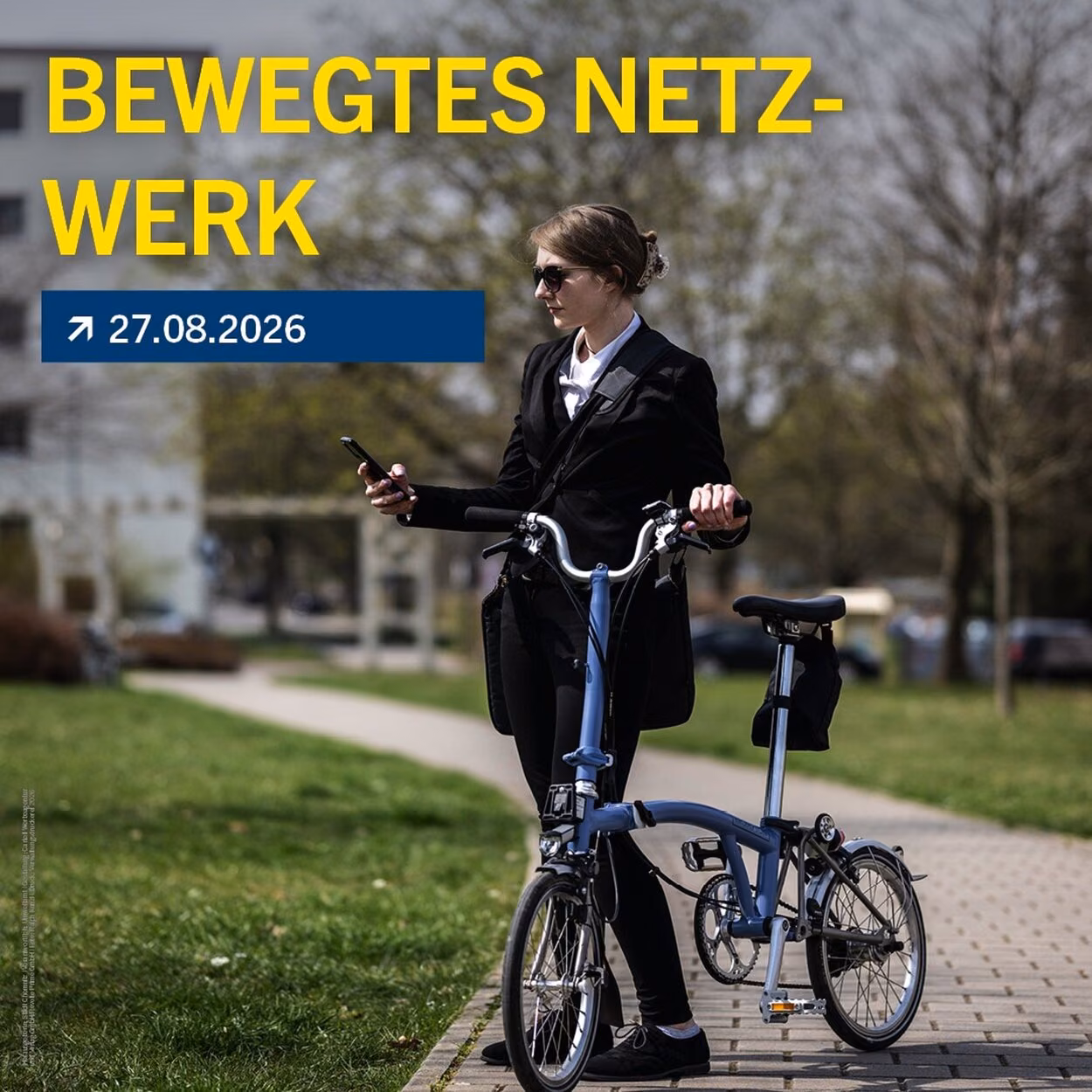 Das Bild zeigt eine Frau in Anzug wie sie auf einem gepflasterten Weg mit ihrem Klappfahrad steht. Ihr Blick ist auf ihr Handy gerichtet. Über ihr stehen in gelber Schrift die Worte "Bewegtes Netzwerk. 27.08.2026"