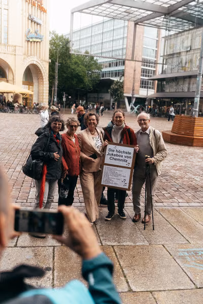Besuchergruppe auf dem Chemnitzer Markt, Stadtmitarbeiterin hält Plakat „Die höchste Jukebox von Chemnitz“, vorne eine Person macht Foto mit Handy.
