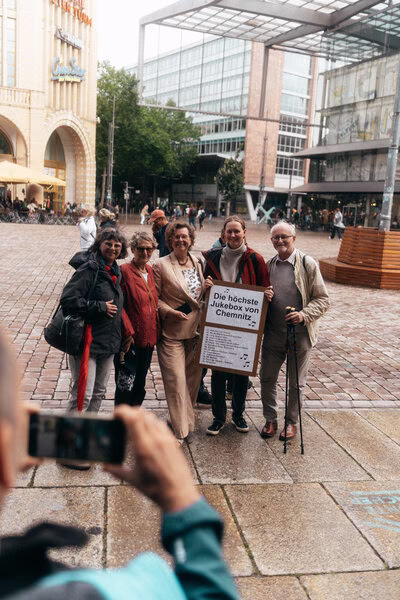 Besuchergruppe auf dem Chemnitzer Markt, Stadtmitarbeiterin hält Plakat „Die höchste Jukebox von Chemnitz“, vorne eine Person macht Foto mit Handy.