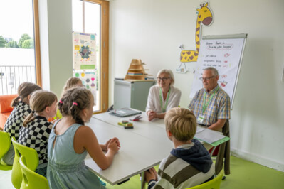 In einem hellen Klassenraum sitzen mehrere Kinder an einem weißen Tisch und hören einer älteren Frau und einem älteren Mann zu, die ihnen gegenüber Platz genommen haben. Die Seniorpartner tragen Namensschilder an grünen Bändern und lächeln freundlich. Im Hintergrund hängt ein Flipchart mit handgeschriebenen Regeln und einer gezeichneten Giraffe an der Wand. Durch das große Fenster fällt Tageslicht in den Raum, während draußen Bäume und ein Gebäude zu sehen sind. Die Atmosphäre wirkt einladend und konzentriert.