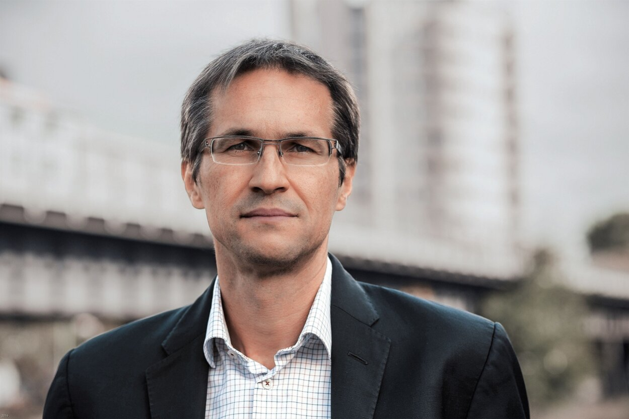 Im Bild ist ein Mann zu sehen, er hat kurze dunkelbraune Haare und trägt eine Brille sowie ein schwarzes Sakko und darunter ein helles Hemd. Er steht im Freien, hinter ihm sind Gebäude in weiterer Entfernung zu erkennen.