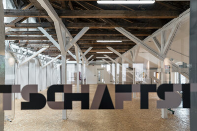 Blick durch eine mit „Stadtwirtschaft“ beschriftete Glastür in einen Dachgeschossraum mit zahlreichen Holzbalken.
