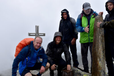 Das Bild zeigt mehrere Personen vor dem Gipfelkreuz. Alle Tragen Regenfeste Wanderkleidung. Die beiden linken Personen hocken. Die linke Person ist Bürgermeister Knut Kunze. Die Person rechts neben ihm ist Bürgermeister Michael Stötzer. Alle Personen schauen freundlich in die Kamera. Im Hintergrund ist nur grauer Nebel zu erkennen.