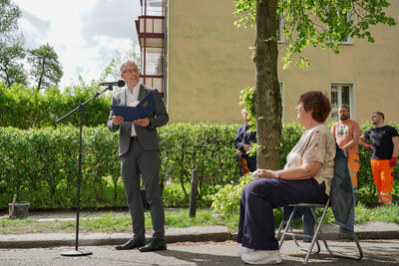Herr Burghart hält eine Rede und lächelt die auf einem Stuhl sitzende Hanna Sachs freundlich an. Im Hintergrund sind ein Musiker und zwei Bauarbeiter zu sehen.