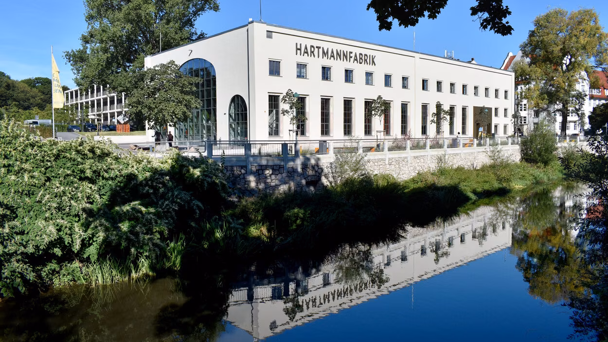 Die Hartmannfabrik spiegelt sich im Fluss Chemnitz, umrahmt von grünen Bäumen.