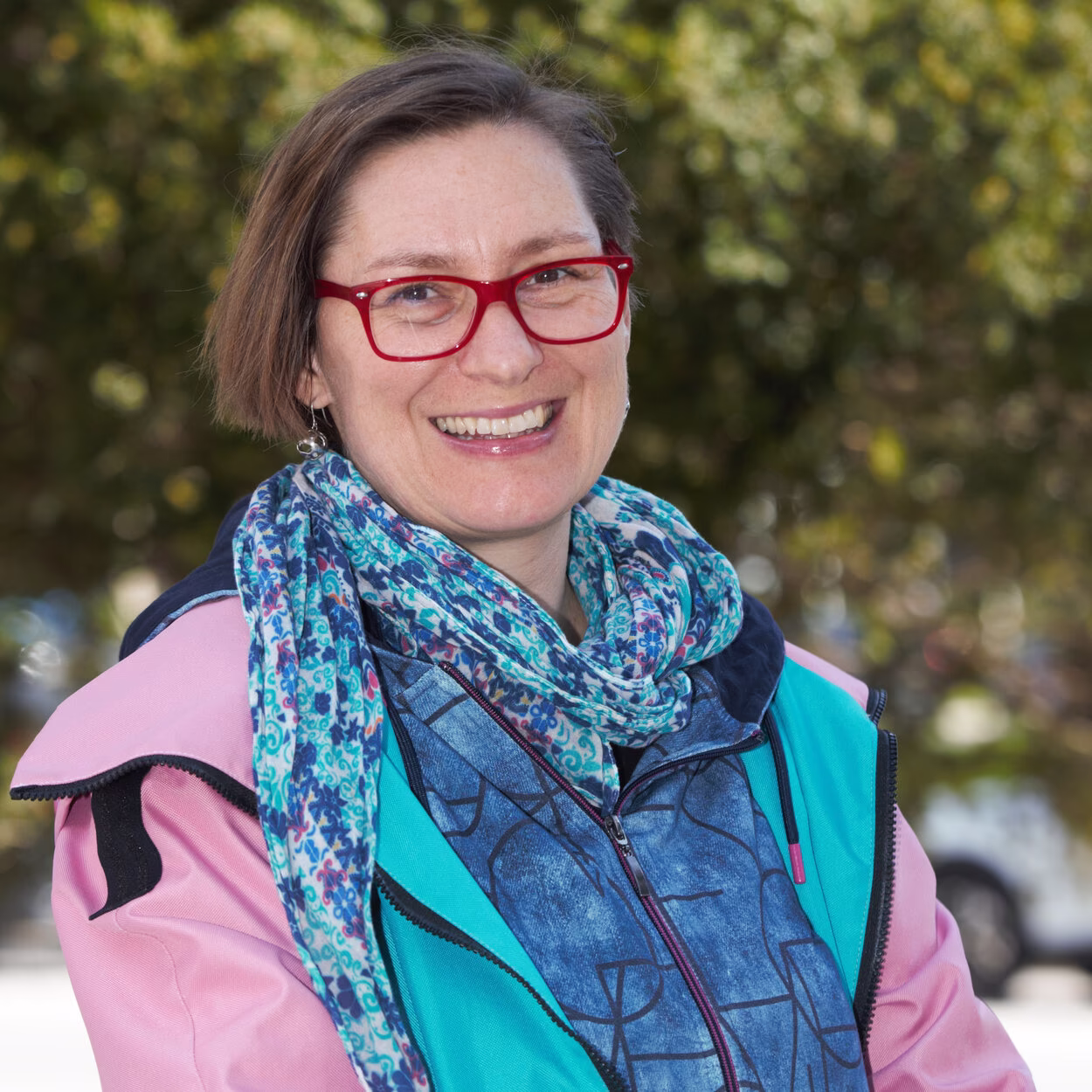 Die Wissenschaftlerin Dr. Mojca Stubelj Ars hat dunkle kurze Haare. Sie trägt eine rote Brille, eine rosa-türkise Jacke und ein großes blaues Tuch. Sie lacht einladend.