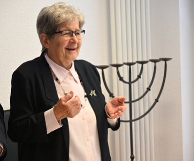 Renate Aris spricht bei einer Feier in der Jüdischen Gemeinde Chemnitz. Sie lächelt, trägt ein dunkles Jackett über einer hellen Bluse und eine Brosche. Im Hintergrund steht ein siebenarmiger Leuchter.