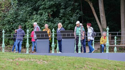 Gäste stehen am historischen Geländer des neu angelegten Aussichtsbalkons unterhalb des Eisenbahnviadukts und lesen die Informationstafeln.