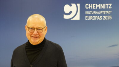 Ulli Engelmann trägt eine runde Brille sowie dunkler Kleidung und steht vor einer großen blauen Wand. Der Hintergrund hat einen sanften Farbverlauf, der an einen Himmel erinnert. Er lächelt freundlich in die Kamera.