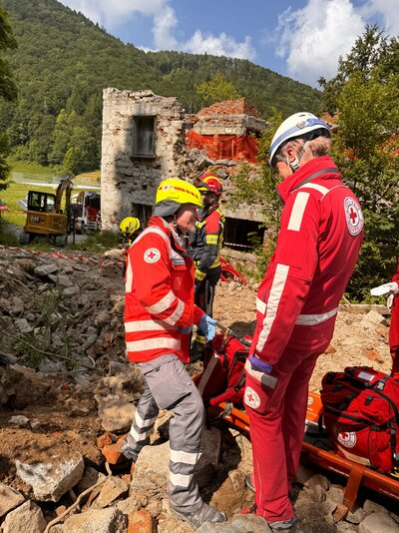 Zwei Männer des Deutschen und des Slowenischen Roten Kreuzes stehen auf einem Schuttberg, im Hintergrund sind eine Ruine und ein Bagger zu sehen.