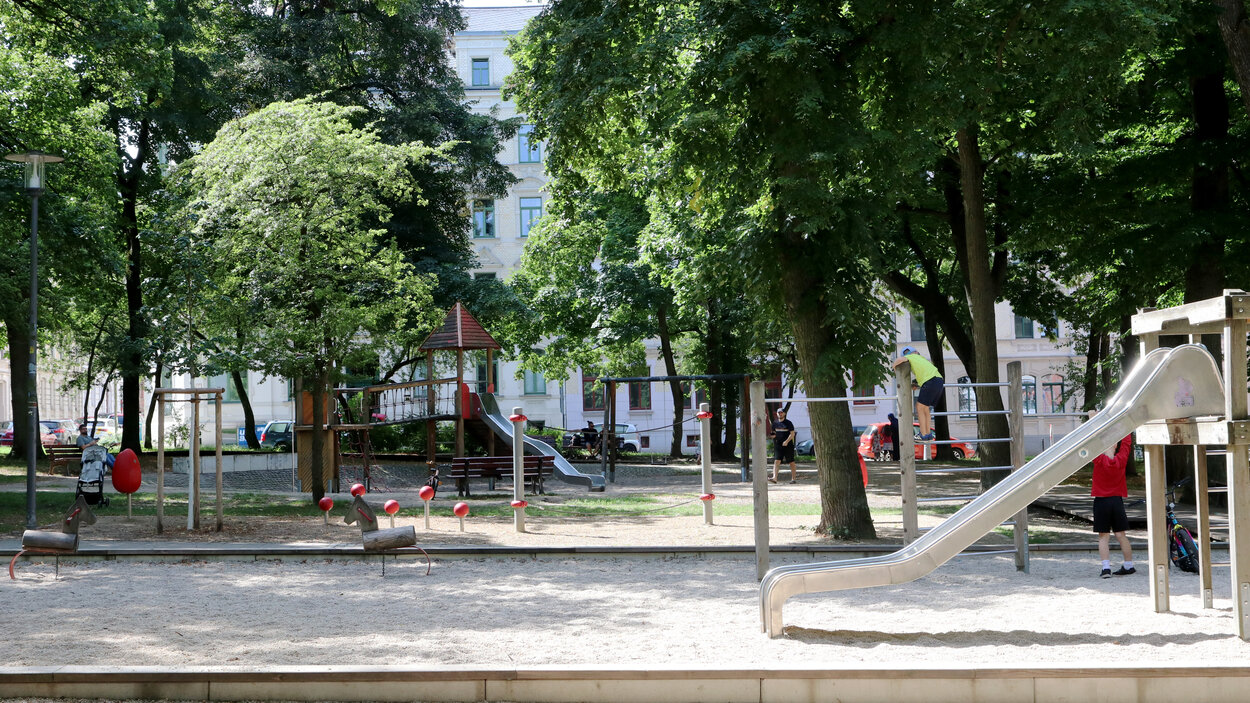 Totale des Lessingplatzes in Chemnitz mit Rutsche auf der rechten Seite, Kiesbett in der Bildmitte und weiteren Spielgeräten im Hintergrund, umstanden von hohen Bäumen.