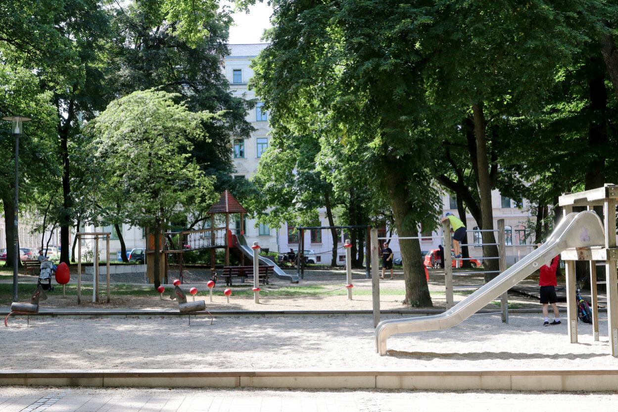 Totale des Lessingplatzes in Chemnitz mit Rutsche auf der rechten Seite, Kiesbett in der Bildmitte und weiteren Spielgeräten im Hintergrund, umstanden von hohen Bäumen.