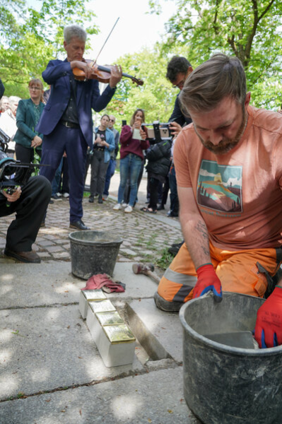 Ein Bauarbeiter verlegt vier Stolpersteine in den Boden; im Hintergrund sind Besucherinnen und Besucher sowie ein Musiker zu sehen, der die Veranstaltung begleitet.