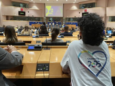 Zasedací místnost z pohledu diváků zezadu. Jeden z diváků má na sobě bílé tričko s potiskem loga Evropského hlavního města kultury Chemnitz 2025 na zádech.