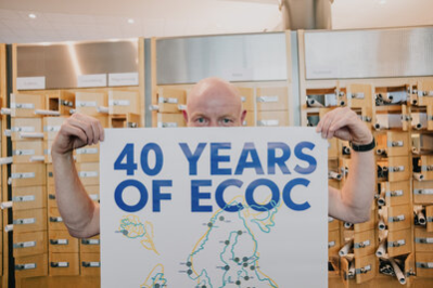 Ein Mann hält ein Poster mit der Aufschrift "40 Years of ECOC". Von ihm sind nur das Gesicht ab den Augen sowie seine Hände und Unterarme zu sehen, der Rest ist vom Plakat verdeckt.