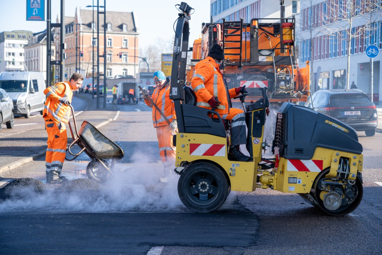 Zwei männliche Mitarbeiter des Bauhofes in oranger Kleidung tragen dampfenden Asphalt auf. Eine Dampfwalze, die ein Mann in Warnkleidung bedient, ebnet den Straßenbelag.
