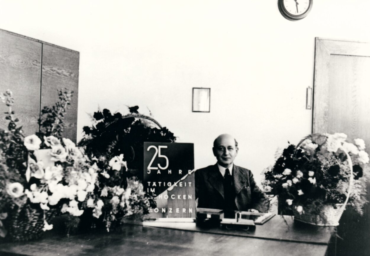 Schwarzweißfotografie von Carl Lewin in einem dunklen Anzug und Krawatte hinter seinem Schreibtisch. Auf dem Schreibtisch sind Blumenbouquets und ein Schild mit der Aufschrift „25 Jahre Tätigkeit Schocken Konzern“ drapiert.