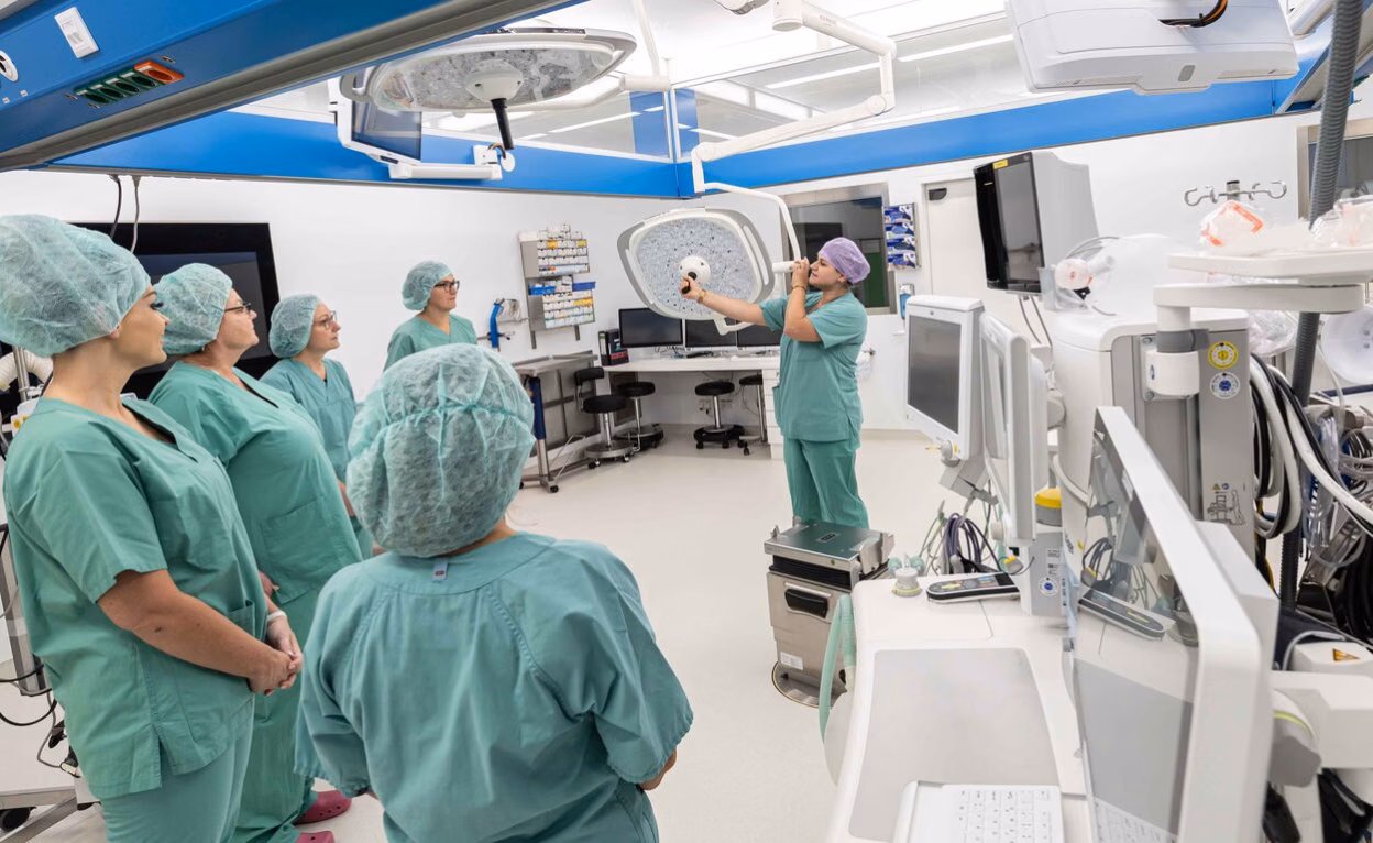 In einem Operationssaal im Klinikum Chemnitz steht eine Gruppe von Menschen in blaugrüner OP-Kleidung und -Hauben. Eine Klinikumsmitarbeiterin demonstriert an einer großen OP-Lampe, wie diese funktioniert. Die Besuchergruppe hört ihr aufmerksam zu.