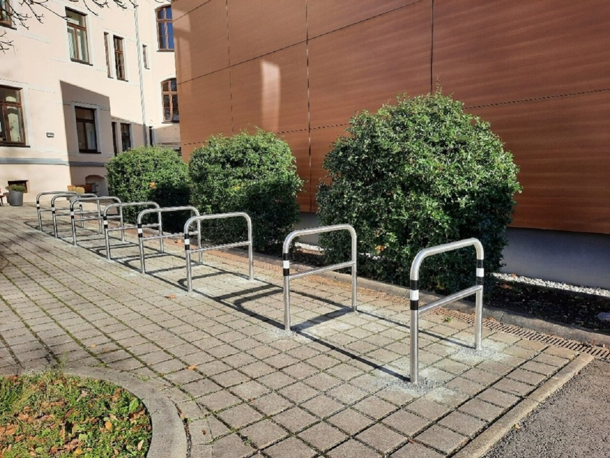 Die Fahrradbügel stehen in einer Reihe nebeneinander auf gepflastertem Boden. Im Hintergrund sind sowohl Gebäude als auch Büsche zu sehen.