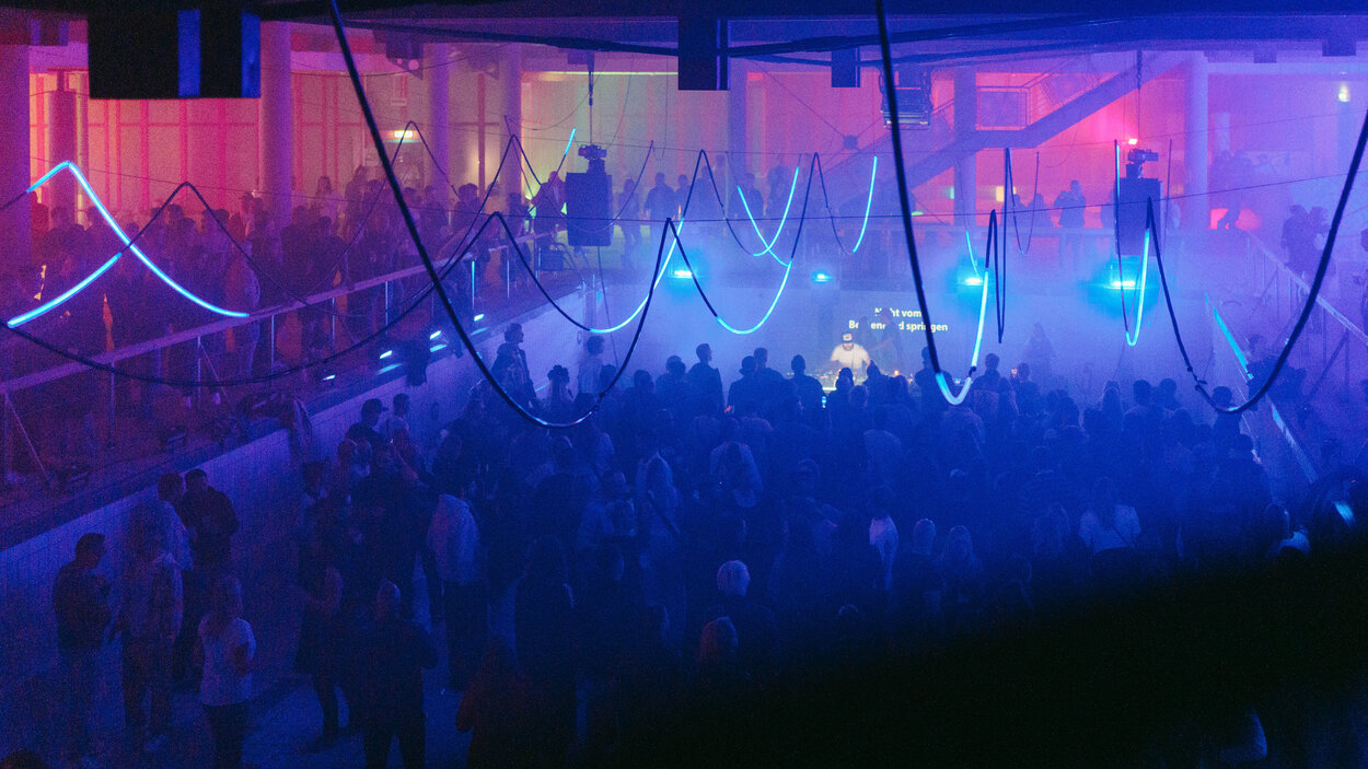 V bývalém plaveckém bazénu tančí na rave party velký dav lidí. Prostor je zalitý sytým modrým, fialovým a růžovým světlem, zatímco halou se prohánějí mlha a paprsky světla. Na stropě a nad tanečním parketem visí zakřivené světelné a dekorativní prvky.