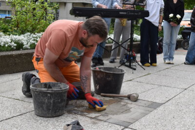 Ein Bauarbeiter wischt nach dem Einlassen in den Boden mit einem Schwamm über den Stolperstein.