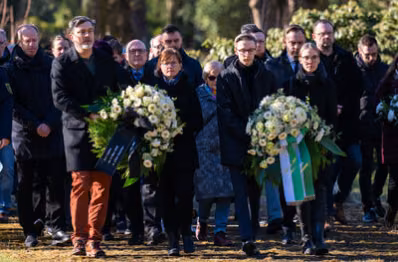 Zu sehen sind die Menschen, die zum Mahnmal laufen. Es werden zwei Kränze nebeneinander getragen.