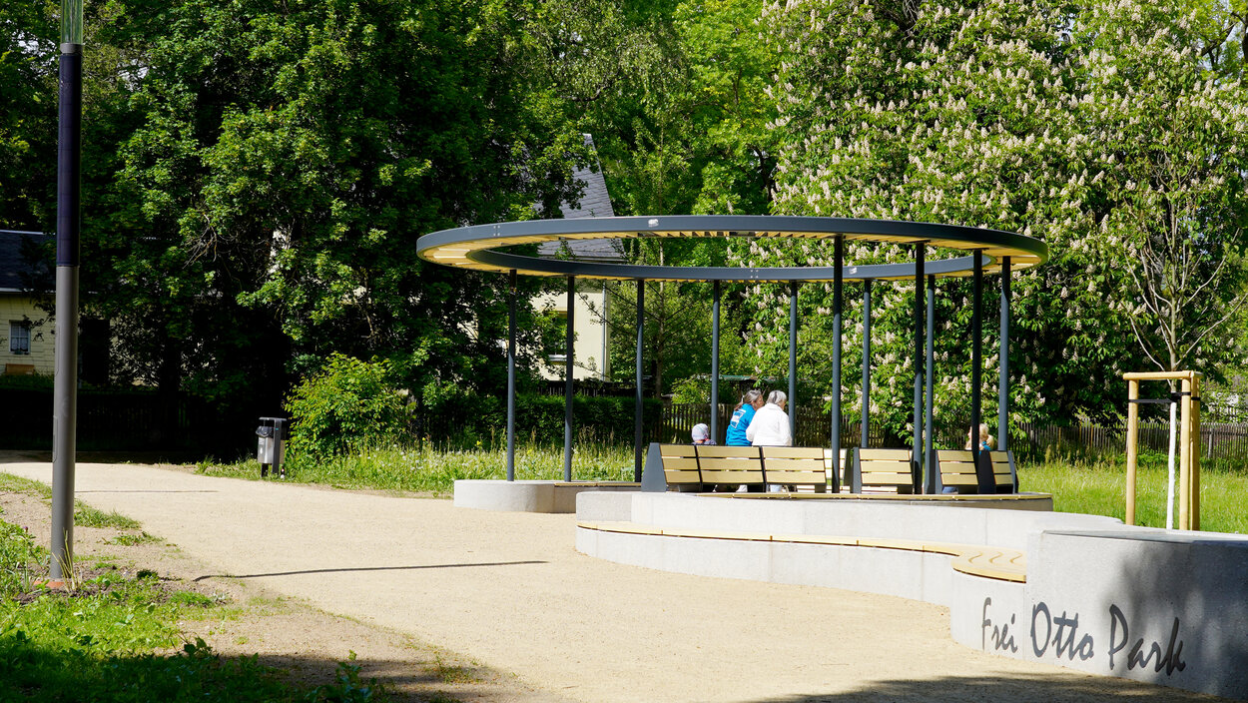 Eine Pergola mit dem Schriftzug "Frei Otto Park" steht entlang eines Weges. Im Zentrum dieser sitzen mehrere Leute und unterhalten sich im Sonnenlicht.