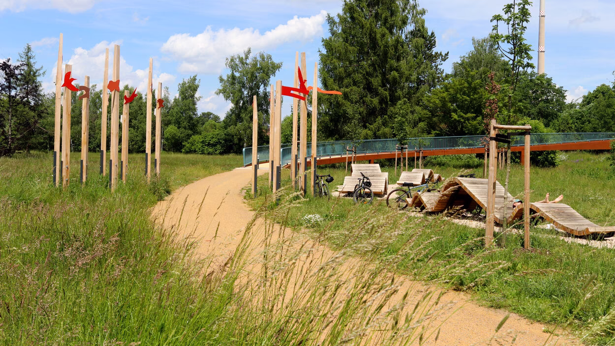 Ein sandgeschlämmter Weg verläuft in einem Bogen durch das Bild in Richtung Brücke; Gräser neigen sich über den Weg, an Holzbalken sind rote Badelemente aus Holz angebracht.