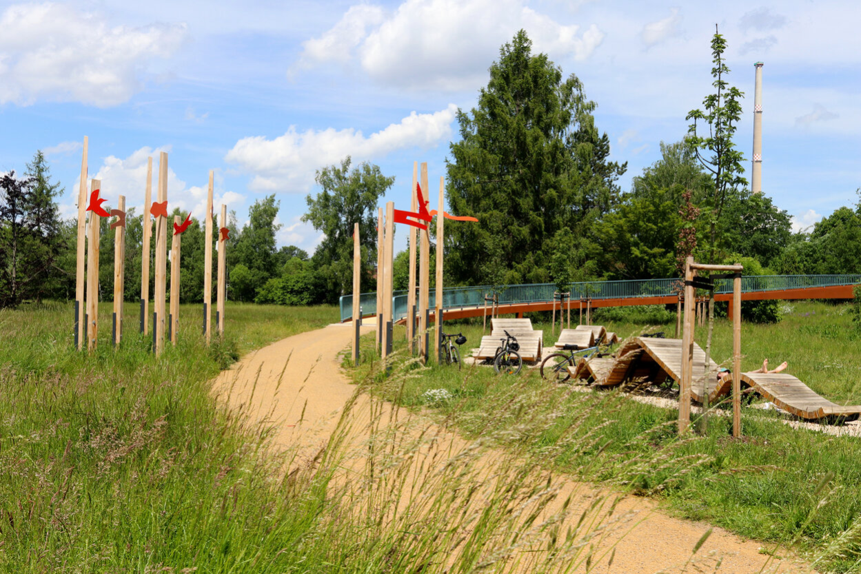 Ein sandgeschlämmter Weg verläuft in einem Bogen durch das Bild in Richtung Brücke; Gräser neigen sich über den Weg, an Holzbalken sind rote Badelemente aus Holz angebracht.