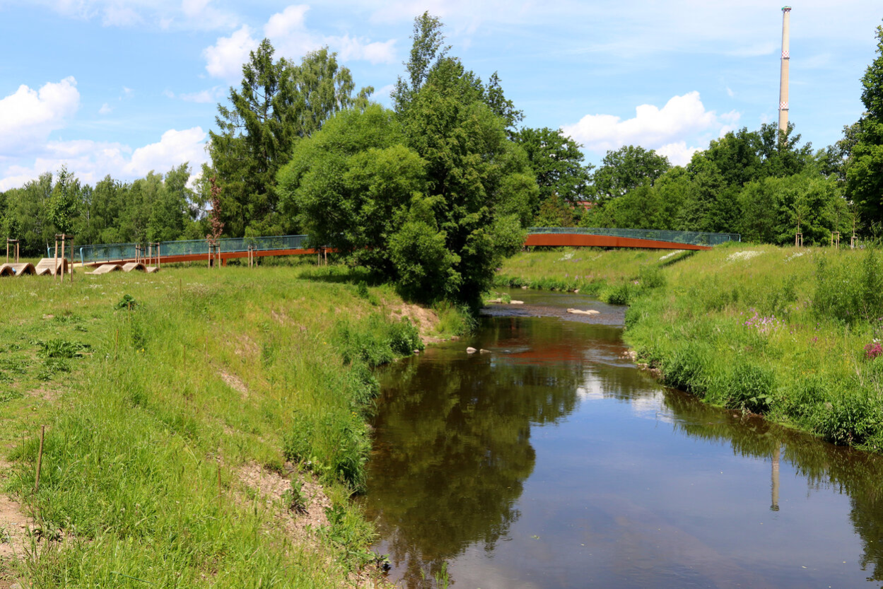 Ein Fluss verläuft von vorn nach hinten durch das Bild, im Hintergrund überspannt ihn eine lange Fußgängerbrücke; umgeben von Wiesen und Bäumen.