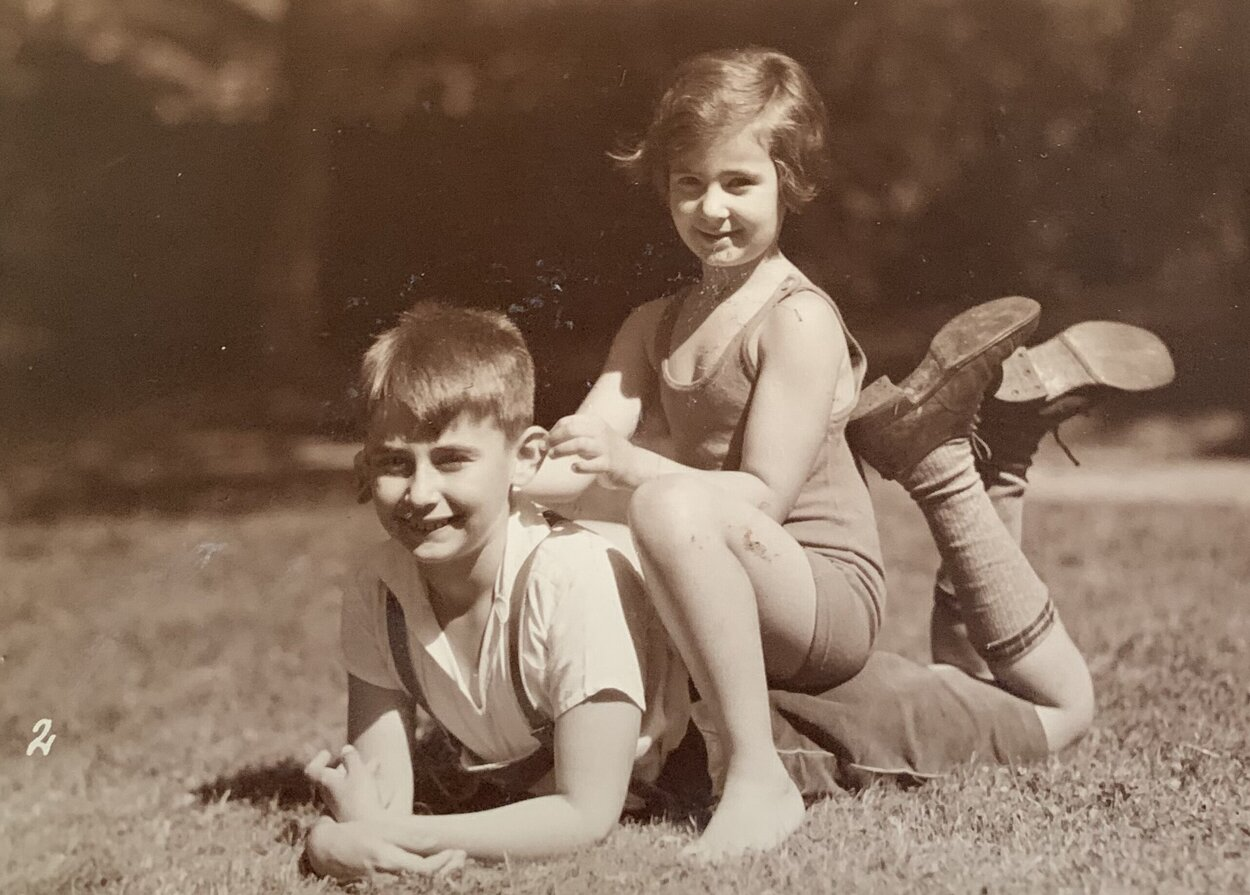 Die Sepia-Aufnahme zeigt die Geschwister Becker als Kinder. Fritz-Edgar liegt auf dem Bauch auf einer Wiese, seine Schwester sitzt auf seinem Rücken, die beiden lächeln in die Kamera. Fritz-Edgar trägt eine Latzhose und darunter ein helles Polohemd sowie Kniestrümpfe und Halbschuhe. Seine Schwester trägt eine kurze Hose und ein ärmelloses Oberteil, sie ist barfuß.