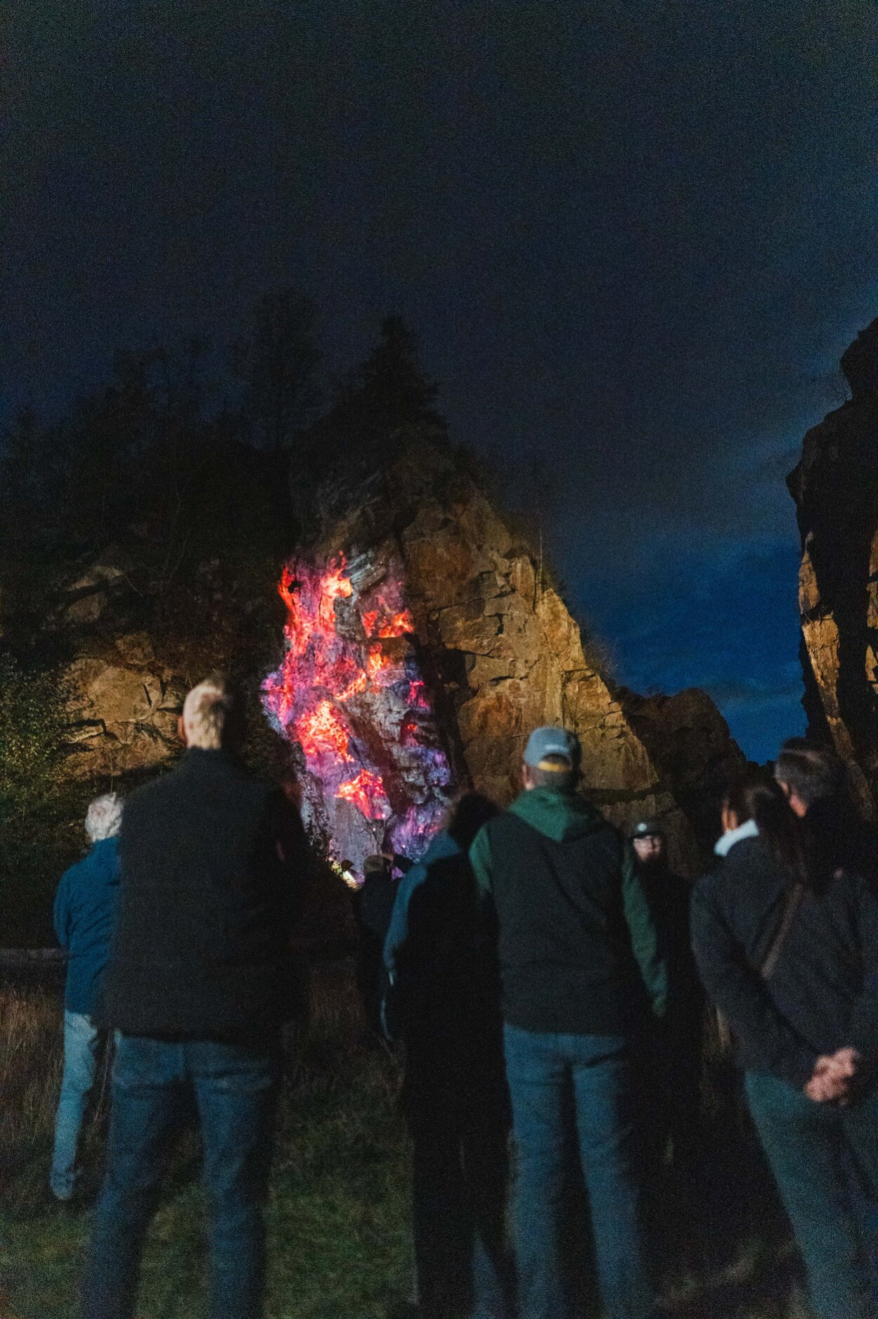 V noční Geyerschen Binge stojí několik lidí před impozantním skalním útvarem. Na tmavé skále září barevná světelná instalace v odstínech červené, růžové, fialové a oranžové. Návštěvníci si osvětlené skály prohlížejí zblízka, zatímco scénu obklopuje temná noční obloha a okolní skalní stěny.
