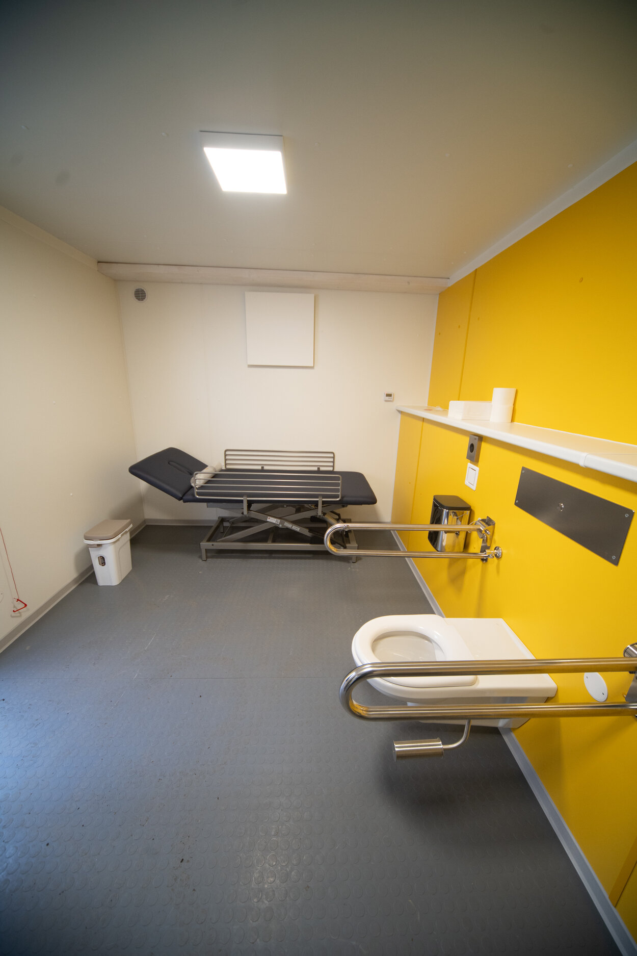The picture shows the interior of the "toilet for all". There is a wide, dark grey lounger on the back wall, with a small grille on either side for protection and to hold on to. The right-hand wall is painted a sunny yellow colour. The low toilet is attached to it, next to which there are also metal handles on both sides to hold on to. There is also a waste bin, an emergency call system with a pull cord and other utensils in the "toilet for all".