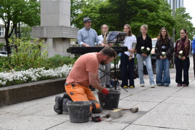 in Bauarbeiter bereitet die Verlegung der Stolpersteine vor; im Hintergrund musizieren Schülerinnen des Georgius-Agricola-Gymnasiums Chemnitz.