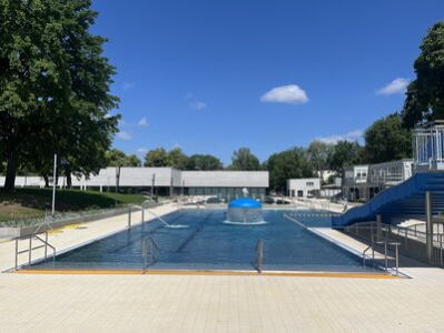 Im Fokus des Bildes steht das Mehrzweckbecken mit Wellenrutsche. Dahinter befindet sich das 50 Meter Schwimmbecken sowie das Gebäude von dem Hallenbad.