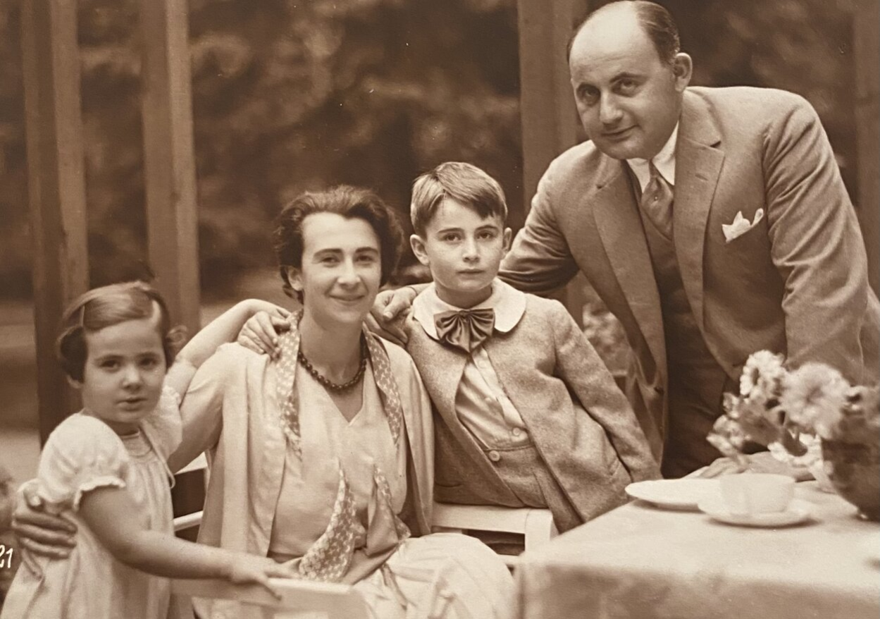 Die Sepia-Aufnahme ist ein Familienfoto der Familie Becker mit Arthur und Charlotte Becker mit ihren Kindern Fritz-Edgar und Liesel. Charlotte Becker sitzt an einer Kaffeetafel, von beiden Seiten haben ihre Kinder den Arm um ihre Schultern gelegt und sie hält ihre Kinder ebenfalls in ihren Armen. Sie lächelt fröhlich in die Kamera. Arthur Becker ist rechts im Bild zu sehen, er steht neben seinem Sohn und hat den Arm um ihn gelegt. Sie sind festlich gekleidet. Auf dem Tisch am rechten unteren Bildrand ist eine Vase mit Blumen zu erkennen sowie ein Kaffeeservice.
