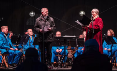 The Very Reverend John Witcombe in schwarzer Kleidung und Nancy Gibson, Leiterin der Städtischen Musikschule, im roten Mantel, stehen auf einer Bühne vor zwei Mikrophonen. Im Hintergrund sitzt die Band GoNG aus Nova Gorica/Gorizia in hellblauen Jacken und Hosen.