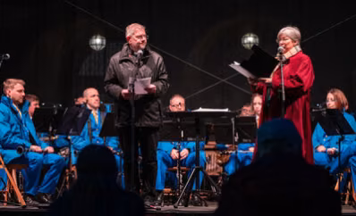 The Very Reverend John Witcombe in schwarzer Kleidung und Nancy Gibson, Leiterin der Städtischen Musikschule, im roten Mantel, stehen auf einer Bühne vor zwei Mikrophonen. Im Hintergrund sitzt die Band GoNG aus Nova Gorica/Gorizia in hellblauen Jacken und Hosen.