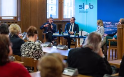 Bei der Podiumsdiskussion sitzt Publikum im Saal. The Very Reverend John Witcombe spricht in ein Handmikrophon. Die anderen Podiumsteilnehmer hören interessiert zu.