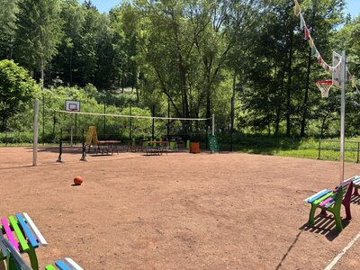 V popředí hřiště na míčové sporty se sítí, v pozadí les. Po stranách jsou barevné lavičky.
