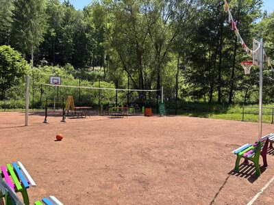 In the foreground a ball sports field with a net, in the background a forest. There are colourful benches at the sides.