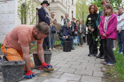 Ein Bauarbeiter reinigt die Stolpersteine mit einem Schwamm von Befestigungsmaterial; im Hintergrund sind Besucherinnen und Besucher sowie Carolin Guttman zu sehen.