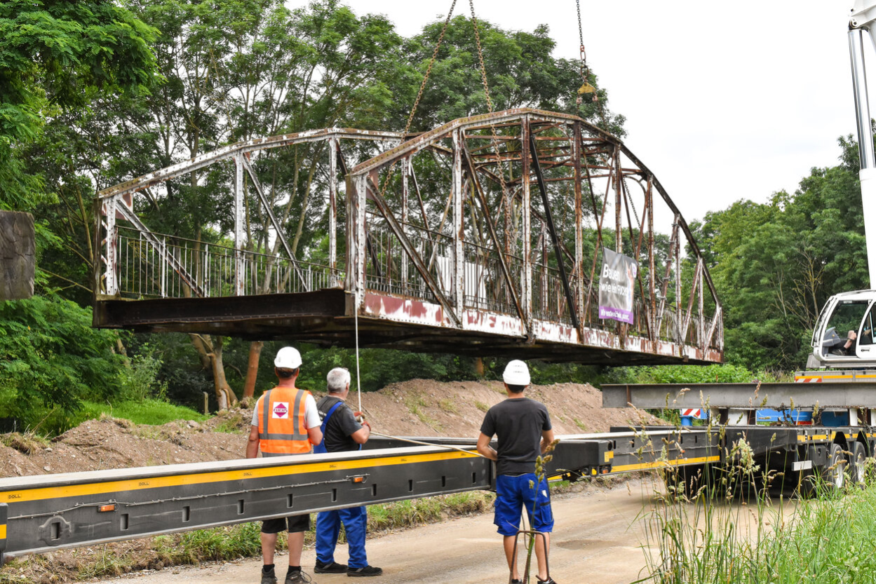 Historische Fischwegbrücke hängt am Kran, drei Arbeiter ziehen sie mit Seilen auf einen Tieflader.
