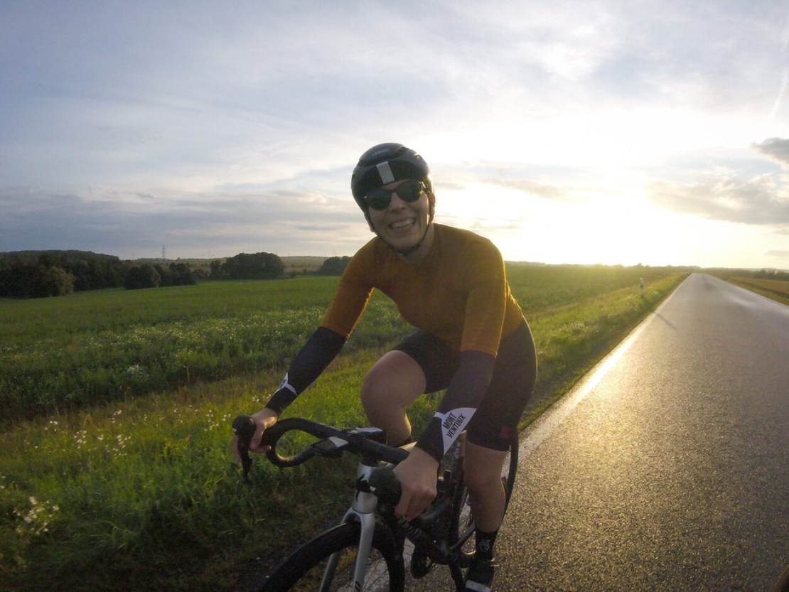 Im Bild ist eine Radfahrerin zu sehen, die auf einer Straße auf den Betrachter zufährt. Sie fährt ein Rennrad mit dem typischen gebogenen Lenkrad. Sie trägt einen Helm und lächelt in die Kamera. Der Himmel ist blau, sonnig und ganz leicht bewölkt. Neben der Straße ist ein Feld mit grüner Bepflanzung.