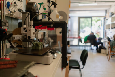3D-Drucker auf einem Tisch im linken Bildbereich, im Hintergrund Menschen an weiteren Tischen.