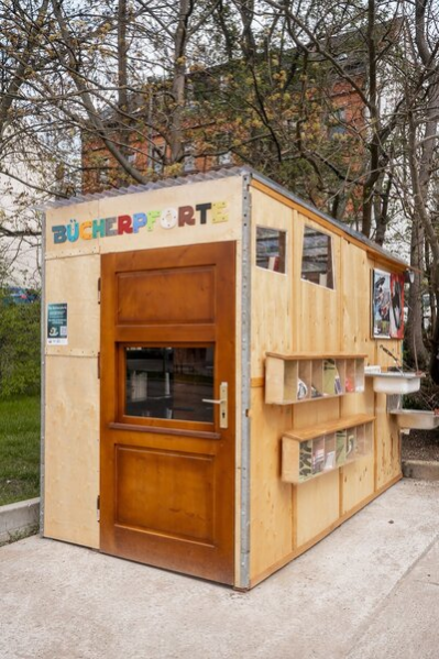 Ein hölzernes Objekt steht auf einem betonierten Boden. Auf diesem ist in bunten Buchstaben "Bücherpforte" geschrieben und eine Tür mit durchsichtigem Fenster führt hinein.