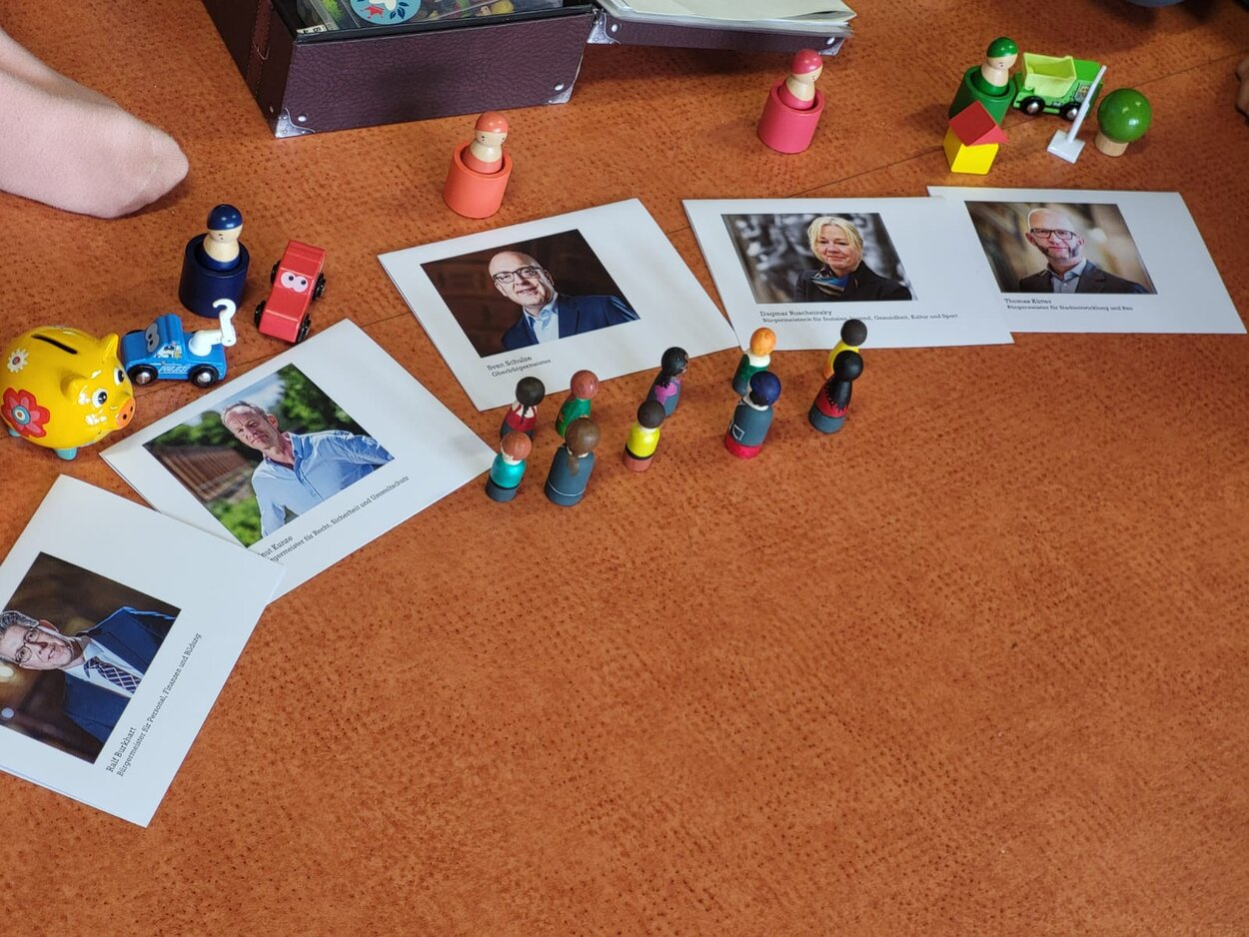 Auf dem Boden liegen Fotos der Bürgermeister der Stadt Chemnitz und verschiedene farbige Holzfiguren.